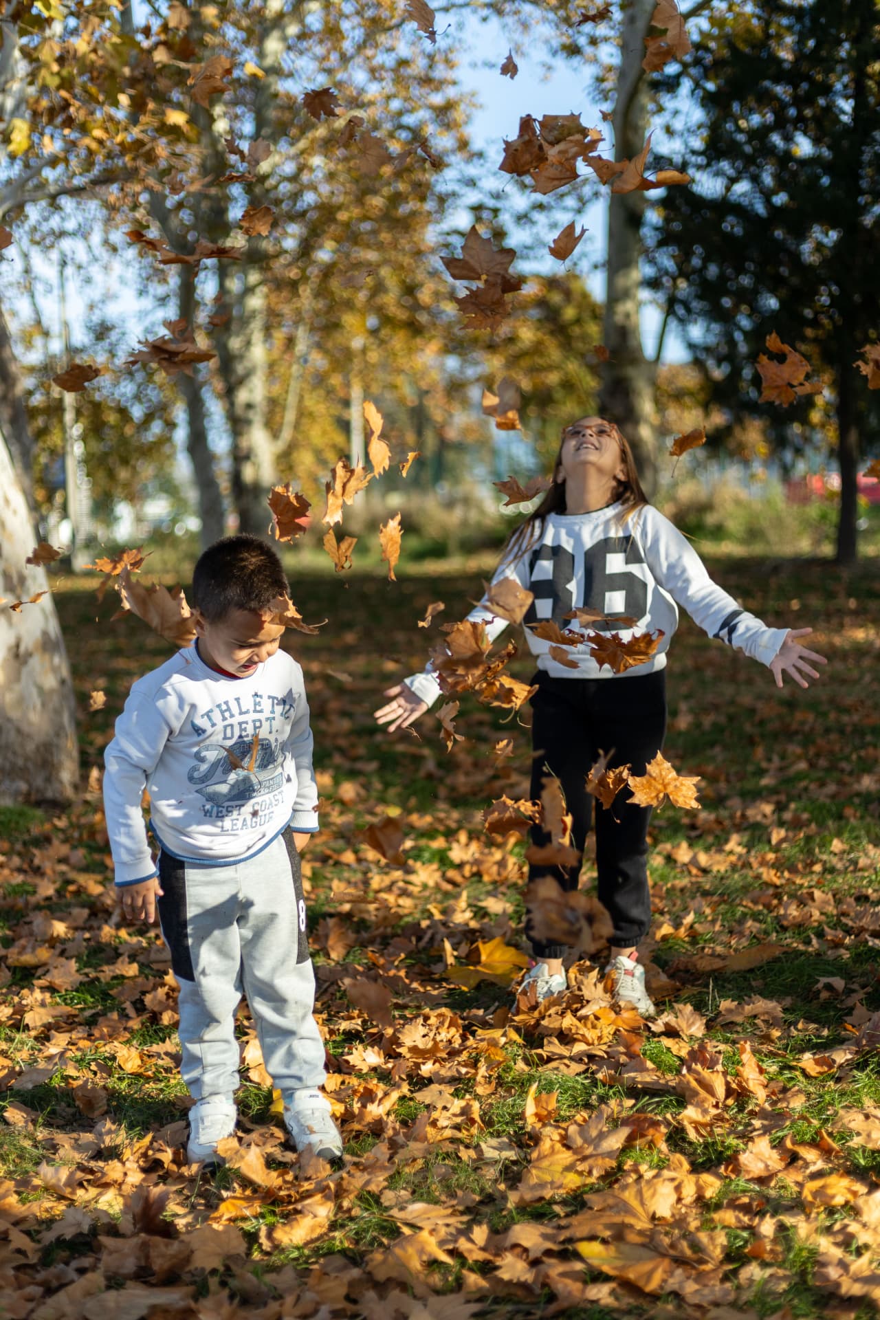 A brother and sister tossing autumn leaves into the air at a park